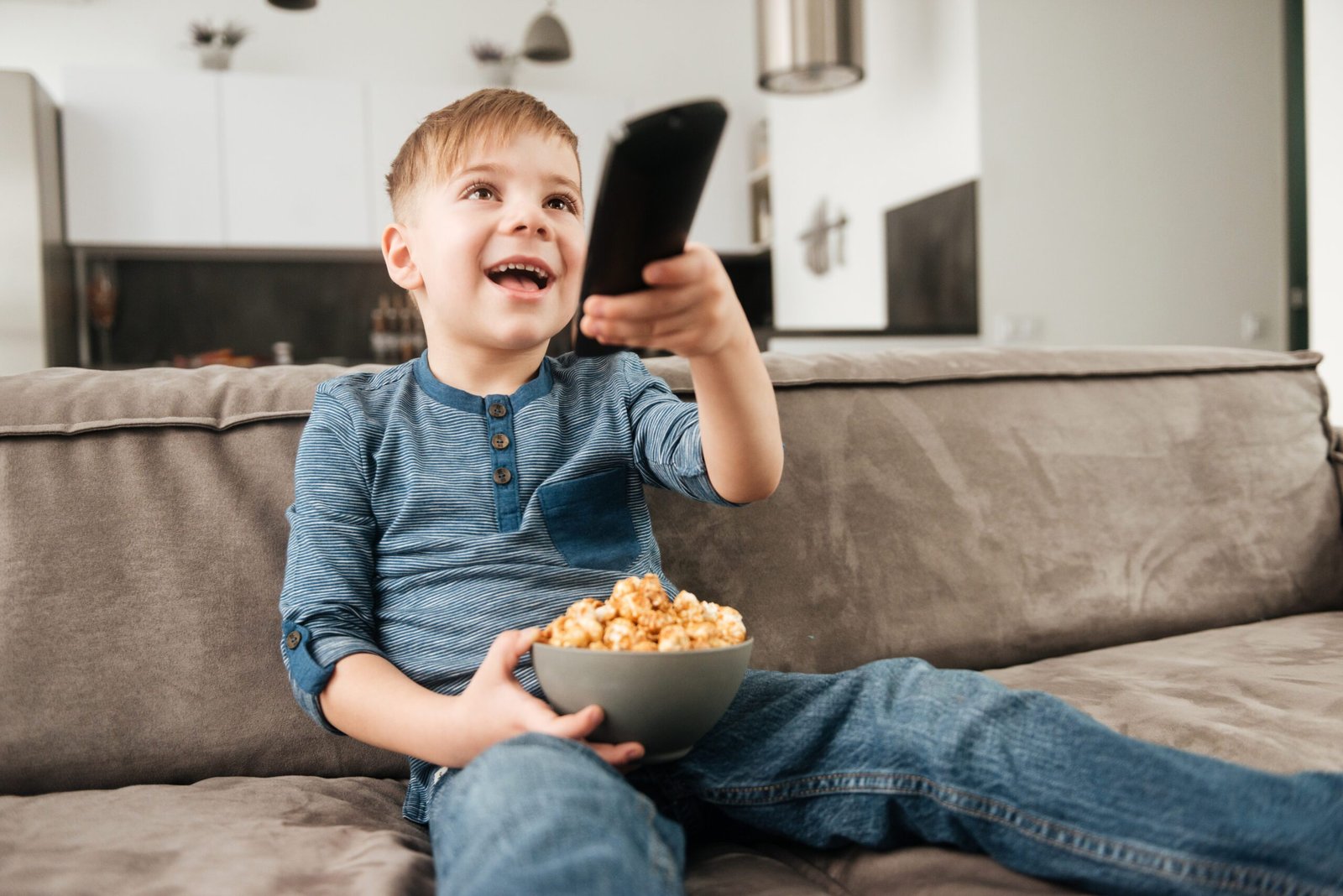 cute boy sitting on sofa holding remote control while watching TV and holding popcorn in hands.