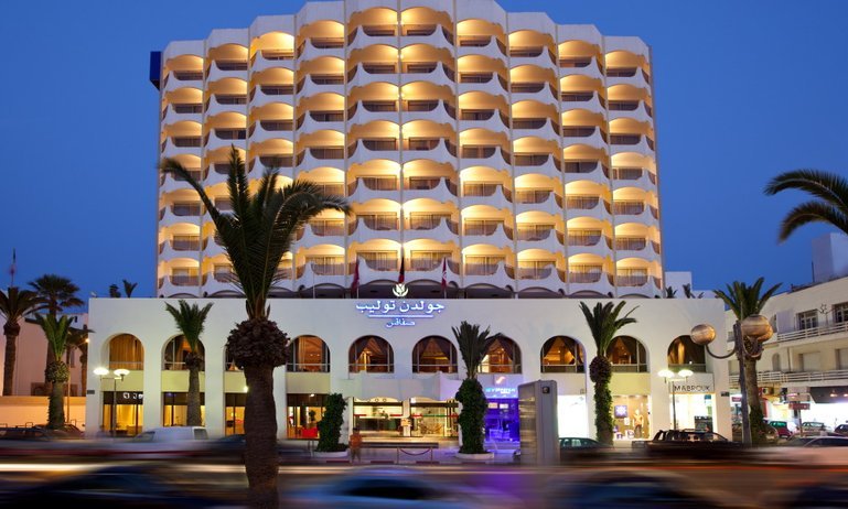 Golden Tulip Sfax hotel illuminated at dusk with palm trees in front.