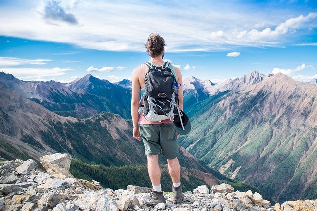 A traveler standing on a rocky mountain peak, overlooking a vast range of mountains under a clear blue sky, embodying the hoptraveler.com travel lifestyle.